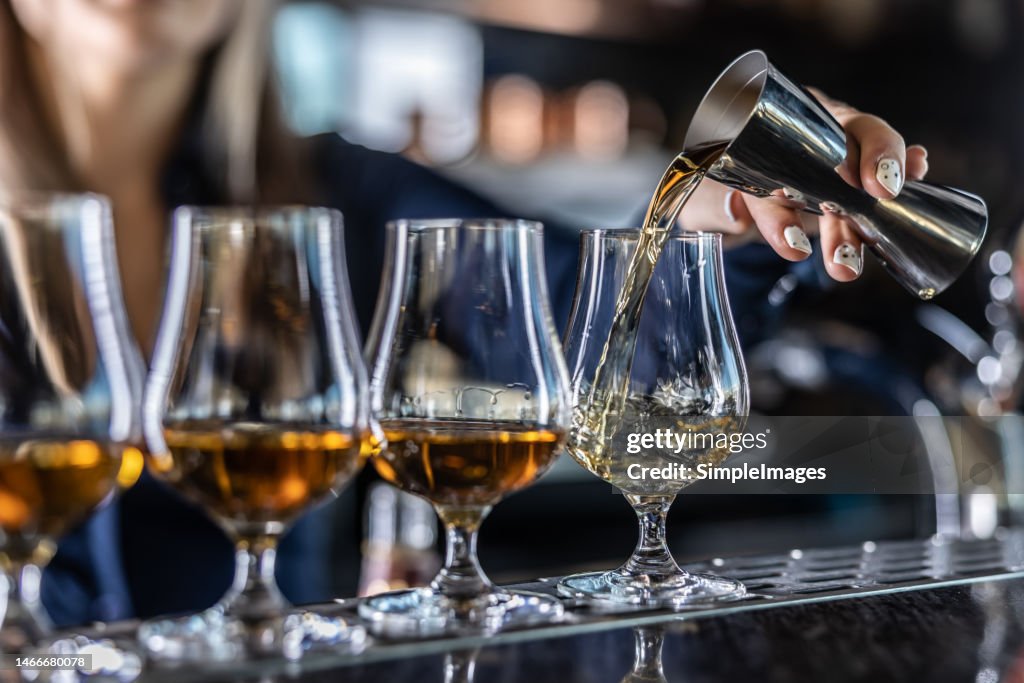 Young female bartender professionally pours rum or brandy from steel jigger into glass at bar.