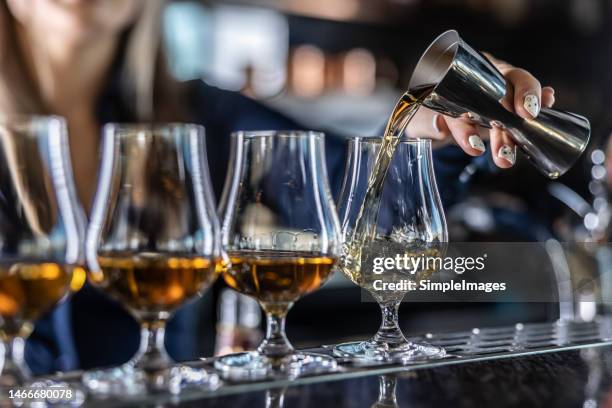 young female bartender professionally pours rum or brandy from steel jigger into glass at bar. - copa fotografías e imágenes de stock