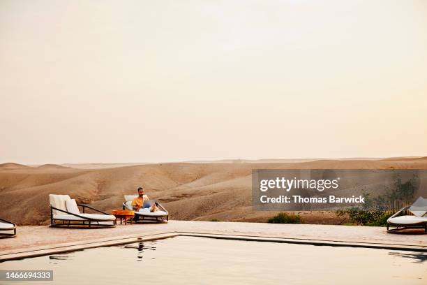 wide shot man sitting beside pool and working on laptop at desert camp - lugar turístico fotografías e imágenes de stock