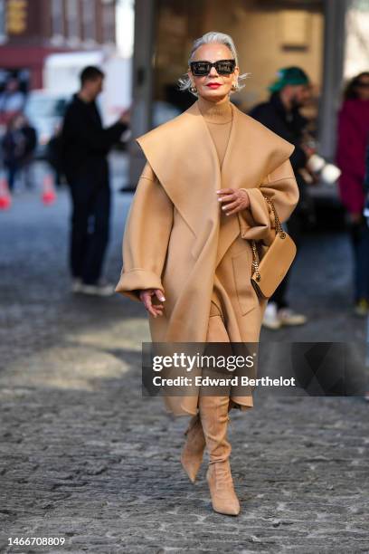 Grece Ghanem wears black sunglasses, gold earrings, a beige turtleneck pullover, a beige long coat with oversized collar, a beige shiny leather...