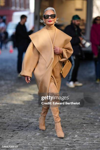 Grece Ghanem wears black sunglasses, gold earrings, a beige turtleneck pullover, a beige long coat with oversized collar, a beige shiny leather...