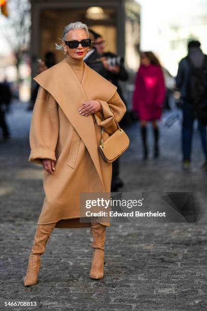 Grece Ghanem wears black sunglasses, gold earrings, a beige turtleneck pullover, a beige long coat with oversized collar, a beige shiny leather...