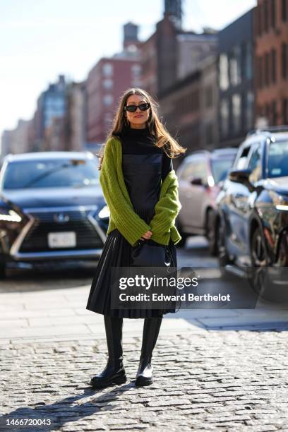 Katerina Dune wears black sunglasses, gold pendant earrings, a black turtleneck / long sleeves t-shirt, a black shiny leather square-neck / pleated...