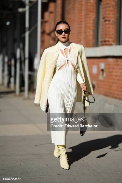 Guest wears black sunglasses, a pale yellow blazer jacket, a white turtleneck / cut-out chest / pleated long dress, a black shiny leather...