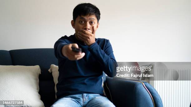 asian man holding remote tv on a couch - awkward stock pictures, royalty-free photos & images