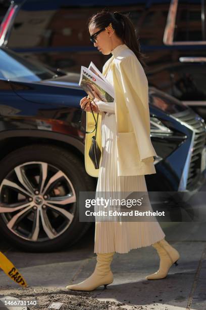 Guest wears black sunglasses, a pale yellow blazer jacket, a white turtleneck / cut-out chest / pleated long dress, a black shiny leather...