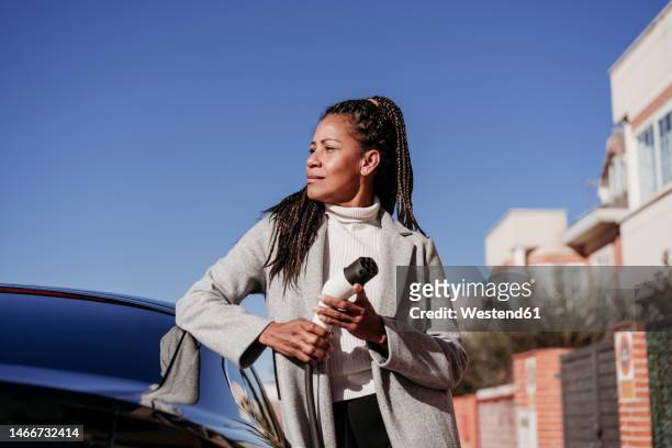 thoughtful woman standing by car and holding electric plug at vehicle charging station - gevlochten haar stockfoto's en -beelden