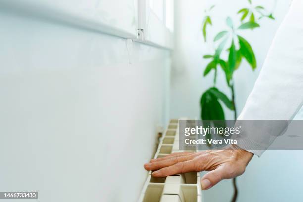 cropped mature woman hand touching home radiator temperature by window - vita domestica fotografías e imágenes de stock
