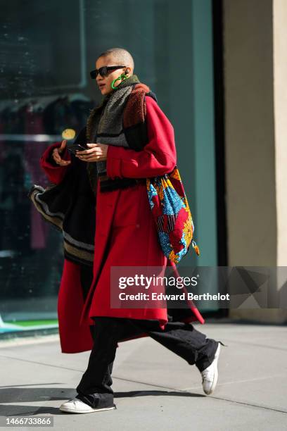 Guest wears black sunglasses, neon green earrings , a black with brown and gray print pattern borders long wool scarf, a red long wool coat, a red...