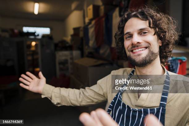 happy young businessman wearing apron inviting in storage room - inviting gesture stock pictures, royalty-free photos & images