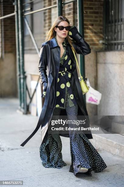Guest wears black vintage sunglasses, a black shiny leather belted long coat, a black with green small polka dots print pattern shirt / long flared...