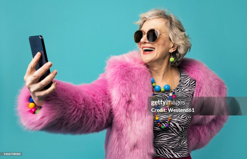 Happy senior woman wearing jewelry taking selfie through smart phone against turquoise background