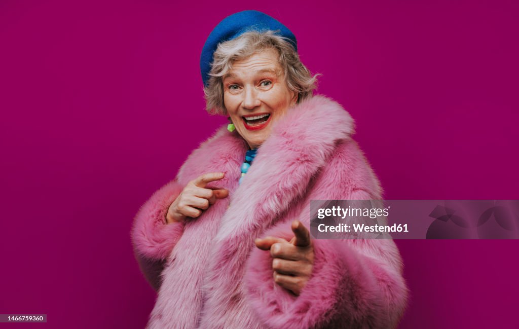 Happy senior woman pink fur coat gesturing against colored background