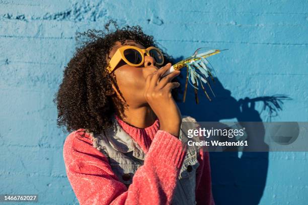 young woman wearing sunglasses and blowing party horn blower - party horn blower imagens e fotografias de stock