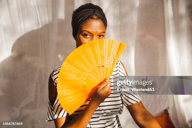 woman covering face with hand fan at home - fächer stock-fotos und bilder