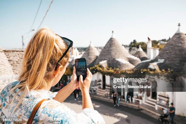 touriste aux cheveux blonds photographiant des maisons trulli avec des toits coniques contre un ciel dégagé à alberobello pendant la journée ensoleillée - trulli photos et images de collection