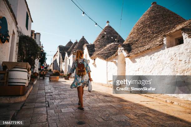 rear view of female tourist with backpack walking in alley amidst trulli houses in a row against clear sky at alberobello - alberobello stock pictures, royalty-free photos & images