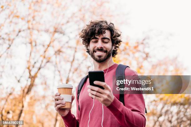 happy man using mobile phone and holding coffee cup standing at footpath - in ear headphones stock pictures, royalty-free photos & images