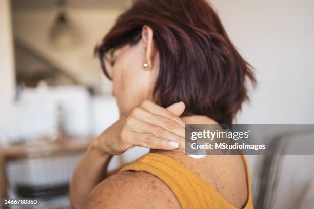 uma mulher latina sênior está sentada no sofá da sala de estar e aplicando um creme hormonal em seu ombro. - hrt pill - fotografias e filmes do acervo