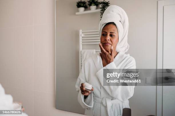 mature woman applying moisturizer on face in bathroom - gezichtscrème stockfoto's en -beelden