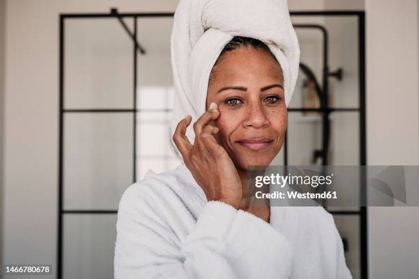 smiling woman applying moisturizer on face in bathroom at home - mature woman - fotografias e filmes do acervo