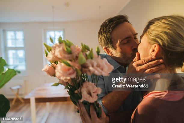 affectionate husband kissing wife holding bunch of flowers at home - woman giving flowers stock-fotos und bilder
