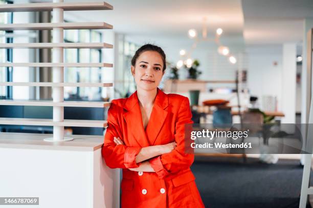 smiling businesswoman with arms crossed standing in office - fato vermelho imagens e fotografias de stock