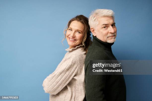 mature man and woman standing back to back against blue background - couple sourire studio photos et images de collection