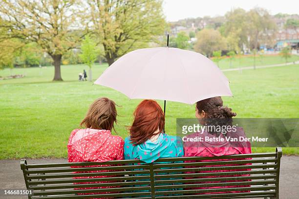 three women sitting under umbrella in park. - sharing umbrella stock pictures, royalty-free photos & images