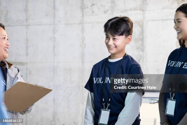 teenagers wearing volunteer shirts are getting assignments from director at food bank while doing charity work together - asian volunteer stock pictures, royalty-free photos & images