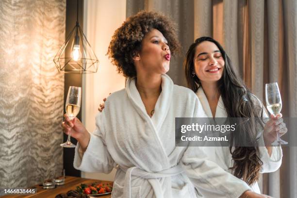 two cheerful diverse friends in bathrobes drinking wine and eating cupcake - ladies night stock pictures, royalty-free photos & images