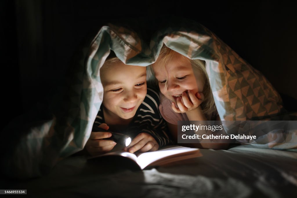 A brother and a sister reading a book with a flashlight under the quilt.