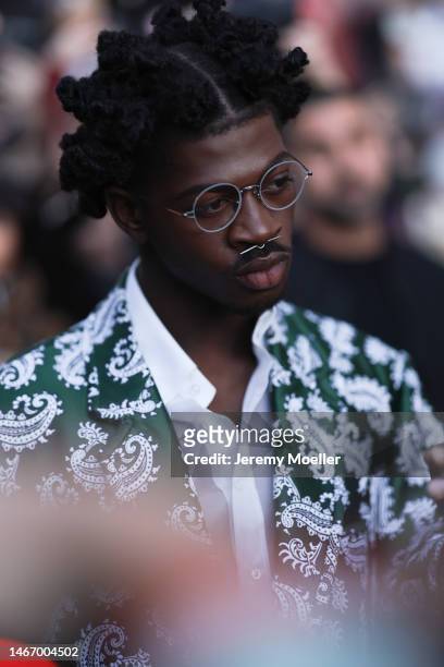 Lil Nas X wears a dark green paisley patterned suit jacket, a white suit shirt and round glasses outside before Thom Browne show on February 14, 2023...