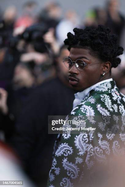 Lil Nas X wears a dark green paisley patterned suit jacket, a white suit shirt and round glasses outside before Thom Browne show on February 14, 2023...