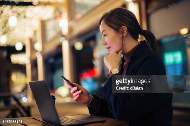 young asian businesswoman log on to her internet banking account on laptop with mobile device on smartphone, sitting outdoors in a side walk cafe at night. privacy protection, internet and mobile security - digital trust stock pictures, royalty-free photos & images