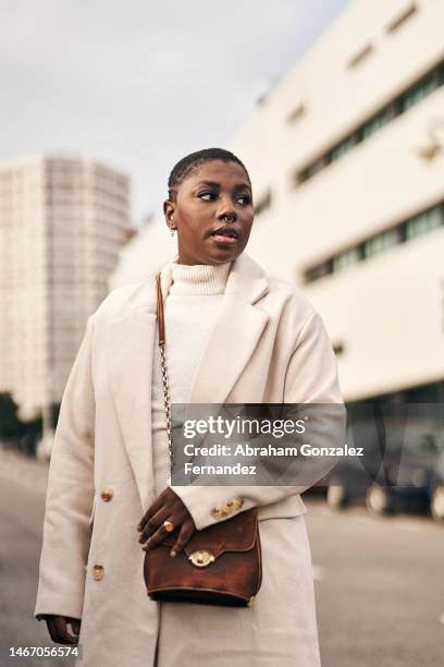 young woman with short hair wearing a coat and carrying her purse standing on the street looking away - handbag stock pictures, royalty-free photos & images