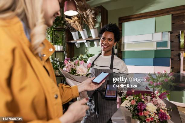 a mature female cashier is smiling while accepting payment by nfc technology at the flower shop. contactless payment concept. - mobile payment stock pictures, royalty-free photos & images