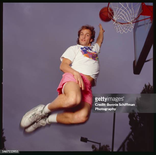 Actor Woody Harrelson is shot from below dunking a basketball in a hoop wearing a white tee and pink shorts in Los Angeles in 1992.