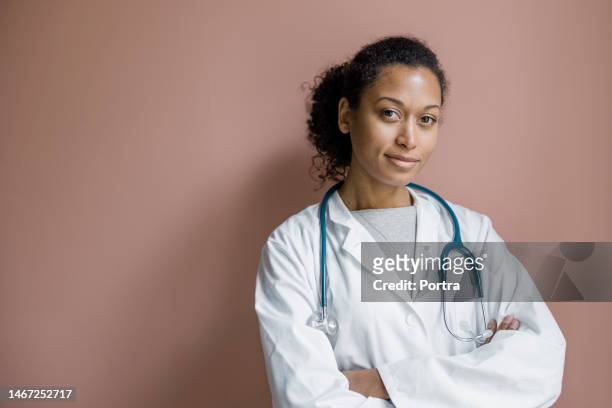 portrait of a confident female doctor standing against brown wall - doctor headshot stock pictures, royalty-free photos & images