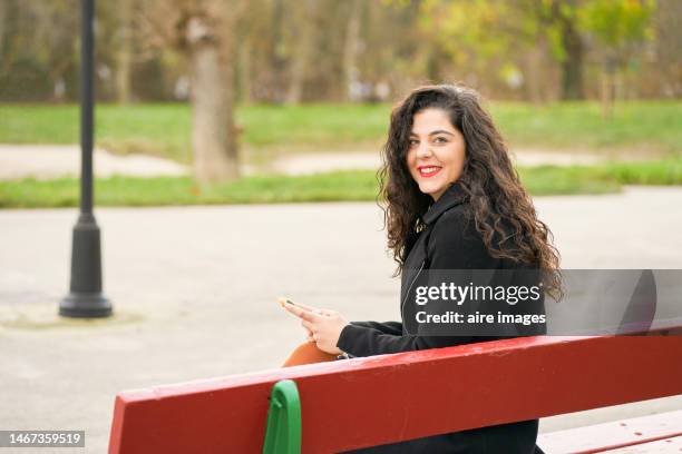 young woman with a smartphone sitting on a red bench in a park. technology concept - lateral portrait looking mobile photos et images de collection