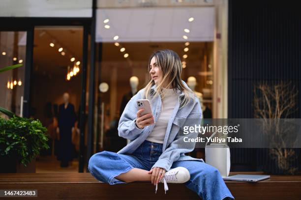 mujer elegante sentada en una playa de madera contra una ventana de una tienda en un centro comercial. - business smartphone happy spring fotografías e imágenes de stock