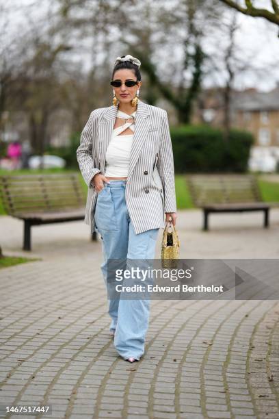 Bettina Looney wears a beige ribbed wool hair elastic, black sunglasses, a white large stones and pendant gold earrings, a gold chain pendant...