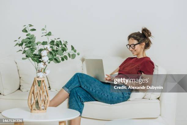 woman sitting on sofa at home using laptop - lesebrille stock-fotos und bilder