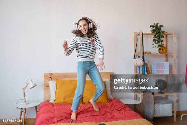 front view of a young girl jumping and dancing on the bed with headphones on. - white bedroom stock pictures, royalty-free photos & images