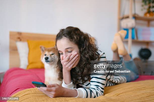 View of a young girl lying face down on her bed, laughing while using her smart phone with her dog beside her.