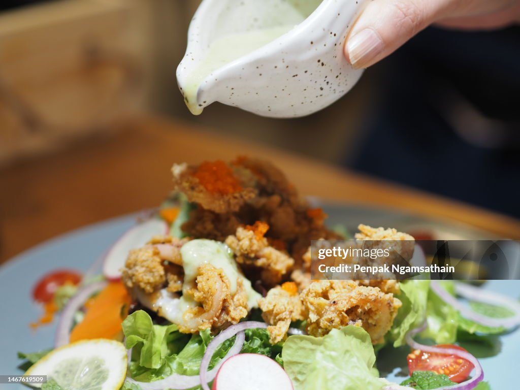 A woman's hand holding a cup pouring sauce on Deep-fried Calamari with Spicy Salad vegetable
