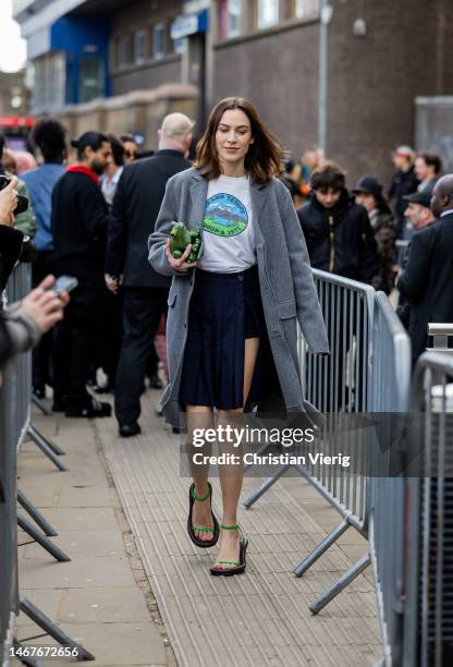 Alexa Chung wears grey coat, navy asymmetric skirt, white top whit graphic print, green bag, sandals outside JW Anderson during London Fashion Week...