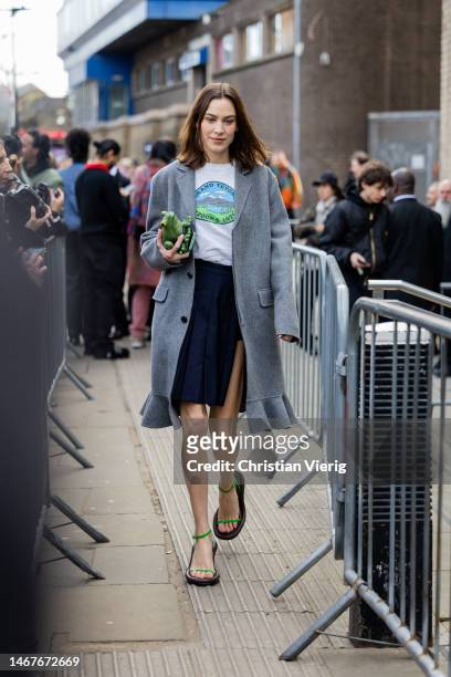 Alexa Chung wears grey coat, navy asymmetric skirt, white top whit graphic print, green bag, sandals outside JW Anderson during London Fashion Week...
