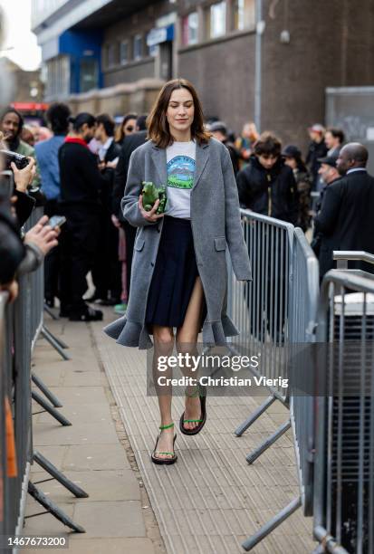 Alexa Chung wears grey coat, navy asymmetric skirt, white top whit graphic print, green bag, sandals outside JW Anderson during London Fashion Week...