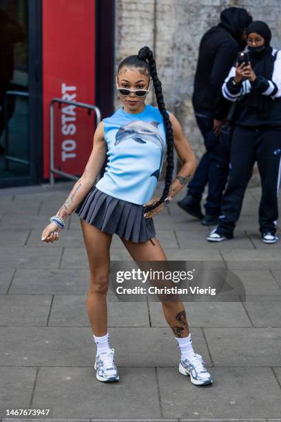 Coi Leray wears grey pleated mini skirt, blue sleeveless top with dolphine print, white socks, sneakers, sunglasses outside JW Anderson during London...